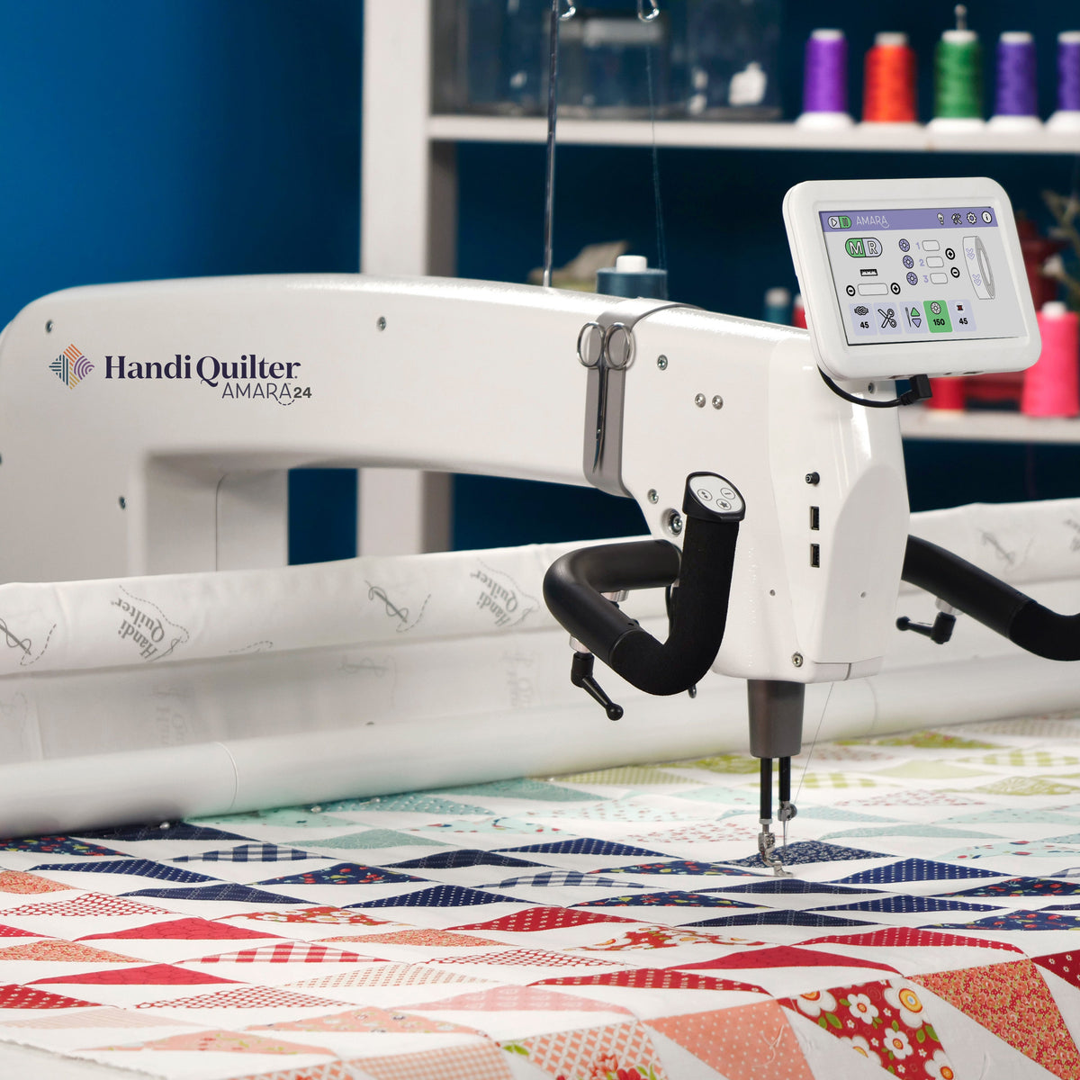 A Handi Quilter Amara 24 longarm quilting machine set up on a table, with a piece of quilted fabric underneath it. The machine has a digital display and is located in a quilting shop with various crafting materials in the background.